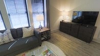 This single-floor space showcases a thoughtfully arranged living area that balances comfort and functionality. The room features warm wood-style plank flooring that runs throughout, providing a cohesive and inviting foundation. Corner windows fitted with horizontal blinds and floor-length curtains allow for versatile natural light control while adding a sense of openness to the space.

The living area is furnished with a charcoal upholstered sofa accented by a geometric-patterned throw pillow, positioned alongside a rustic wood and metal end table with a lower storage shelf. A floor lamp and a table lamp with a chrome base contribute to the warm, ambient lighting that fills the room. A plush area rug anchors the seating arrangement, adding texture and softness underfoot.

Opposite the seating area, a dark wood media console houses a flat-screen television, offering a practical entertainment focal point. The neutral wall tones complement the darker furnishings, creating a balanced and calming atmosphere. Overall, the space presents a well-lit, functional layout suited for relaxed everyday living.
