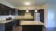 This single-floor space showcases a thoughtfully designed kitchen as its centerpiece. The kitchen is outfitted with rich, dark espresso-toned cabinetry that extends to the upper and lower walls, providing generous storage throughout. Light-toned granite countertops with natural veining offer a striking contrast against the dark cabinetry, creating a visually balanced aesthetic.

A central island, also topped with matching granite, serves as both a workspace and a gathering point, enhancing the kitchen's functionality. Overhead, two pendant lights hang above the island, complementing a flush-mount ceiling fixture that ensures the space is well-lit. The stainless steel appliance suite — including a refrigerator, range, dishwasher, and microwave — adds a modern, cohesive finish to the design.

Dark hardwood-style flooring runs throughout, tying together the deep tones of the cabinetry and grounding the overall look. The neutral, light-colored walls keep the space feeling open and bright despite the bold cabinetry choices. The kitchen's layout provides ample prep space and a functional flow, making it both practical and aesthetically refined.
