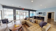 This single-floor community clubroom is designed as a contemporary, open-concept gathering space that seamlessly blends a lounge area with a fully equipped kitchen and bar. The space is anchored by rich wood-look flooring that flows throughout, complementing a carefully curated color palette of cool blues, charcoal gray, and crisp white.

The lounge area is a standout feature, centered around a large sectional sofa with decorative throw pillows and a round wooden coffee table. A dramatic three-dimensional geometric accent wall in white serves as a striking backdrop for a wall-mounted flat-screen television, while an additional TV on the opposite wall ensures visibility from multiple seating areas. A sleek media console and accent chair complete the living space. Floor-to-ceiling glass doors and large windows flood the room with natural light and provide direct views of the outdoor pool area.

Transitioning into the kitchen and bar zone, a spacious white quartz island with bar-height seating takes center stage. Dark navy cabinetry, a stainless steel refrigerator, and an eye-catching blue mosaic tile feature wall add depth and character. Decorative wall-mounted shelving units and curated accessories lend an intentional, styled aesthetic throughout. Additional doorways lead to adjacent utility or support rooms, completing this well-appointed communal space.