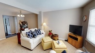 This two-floor space offers a comfortable and inviting atmosphere, with the living area showcasing a well-appointed layout designed for casual relaxation. The living room features neutral beige walls paired with light carpet flooring, creating a warm and understated aesthetic throughout. A textured ceiling adds subtle character to the room.

The seating area is anchored by a white upholstered loveseat accented with bold navy blue patterned throw pillows, providing a striking contrast against the soft, muted backdrop. Yellow tufted ottomans serve as both a coffee table and additional seating, adding a cheerful pop of color and functional flexibility to the space. A wooden media console positioned along one wall accommodates a flat-screen television, making it a practical entertainment area.

Ambient lighting is provided by a classic floor lamp, supplementing the overhead chandelier-style fixture visible in the adjacent area. The open sight line between spaces enhances the sense of flow and connectivity within the home. Overall, the living area exudes a cozy, lived-in charm, balancing comfort and functionality across its two levels.