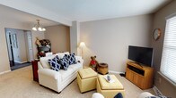 This two-floor space offers a comfortable and inviting atmosphere, with the living area showcasing a well-appointed layout designed for casual relaxation. The living room features neutral beige walls paired with light carpet flooring, creating a warm and understated aesthetic throughout. A textured ceiling adds subtle character to the room.

The seating area is anchored by a white upholstered loveseat accented with bold navy blue patterned throw pillows, providing a striking contrast against the soft, muted backdrop. Yellow tufted ottomans serve as both a coffee table and additional seating, adding a cheerful pop of color and functional flexibility to the space. A wooden media console positioned along one wall accommodates a flat-screen television, making it a practical entertainment area.

Ambient lighting is provided by a classic floor lamp, supplementing the overhead chandelier-style fixture visible in the adjacent area. The open sight line between spaces enhances the sense of flow and connectivity within the home. Overall, the living area exudes a cozy, lived-in charm, balancing comfort and functionality across its two levels.