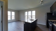 Located at 3610 Dillon St., unit 211, this single-floor space showcases a clean, contemporary design with an open-concept living and kitchen area. The kitchen features a striking dark-cabineted island with a light-toned countertop and an undermount sink equipped with a modern pull-down faucet, offering both functionality and visual contrast. Track lighting overhead provides focused illumination, complementing the natural light that floods the space through multiple large windows along the walls. The windows frame outdoor views while brightening the neutral gray walls and enhancing the airy atmosphere throughout. Rich, dark hardwood-style flooring runs consistently across the entire space, grounding the open layout and adding warmth to the modern aesthetic. The generous open floor plan connects the kitchen island seamlessly to the living area, creating a versatile and inviting environment well-suited for both everyday living and entertaining. The overall ambiance is modern and minimalist, with a carefully balanced palette of dark and light tones that gives the space a polished, well-designed character.