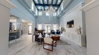 This elegantly designed single-floor community clubhouse or leasing lounge offers a striking first impression with its dramatically tall ceilings accented by bold teal-blue exposed beams. A cascading, sculptural chandelier serves as the focal centerpiece of the expansive main lounge area, drawing the eye upward and amplifying the sense of grandeur. Light-toned wood-look flooring runs throughout the space, complementing the soft blue-gray walls and crisp white wainscoting and shiplap wall panels that lend a coastal, contemporary character.

The main lounge is generously furnished with plush seating arrangements, including velvet accent chairs in deep navy and modern upholstered sofas, creating comfortable gathering areas bathed in natural light from large windows and glass entry doors. Views through the doors reveal an outdoor pool courtyard, seamlessly connecting the interior to outdoor amenities.

Multiple seating vignettes are thoughtfully arranged throughout, including a cozy lounge corner featuring a flat-screen TV mounted on a shiplap accent wall, flanked by sculptural décor and a geometric-patterned media console. An adjacent refreshment bar area features a granite-topped counter, hexagonal tile backsplash in blue, gold, and gray tones, modern wire barstools, and a coffee station, making it an inviting spot for casual gatherings. The overall ambiance is polished, airy, and welcoming.
