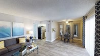 This single-floor residence offers an open-concept layout that seamlessly connects the living, dining, and kitchen areas, creating a spacious and functional flow throughout the home. The living area features wood-style laminate flooring that extends across the main living spaces, providing a clean and contemporary aesthetic. A glass-top coffee table and a comfortable sofa anchor the living room, which benefits from an airy, open feel.

The kitchen is positioned just off the main living area and is equipped with stainless steel appliances, including a full-size refrigerator, and features light-toned cabinetry. A half-wall partition subtly defines the transition between the kitchen and the adjacent dining area, adding architectural interest without closing off the space.

The dining area accommodates a glass-top dining table with seating for multiple guests and is enhanced by a ceiling fan with a light fixture, contributing to both comfort and ambiance. Mirrored wall panels in the dining area reflect light and give the impression of a larger, brighter space.

Throughout, the neutral wall tones and textured ceilings create a warm, inviting atmosphere. A hallway visible from the living room leads to additional rooms, completing this well-organized and thoughtfully designed single-level home.
