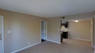 This single-floor residence offers a straightforward and functional layout designed to maximize usability. The living area is finished with light-toned ceramic tile flooring that extends seamlessly throughout the space, creating a cohesive and easy-to-maintain surface underfoot. Walls are painted in a warm neutral beige, complemented by clean white baseboards that provide a crisp contrast.

The open layout allows for a clear sightline from the living area into the kitchen, which features dark espresso-toned cabinetry that adds a bold contrast against the lighter surroundings. The kitchen is equipped with a sink set into a light-colored countertop and includes overhead cabinetry for ample storage. A ceiling-mounted ventilation vent is positioned nearby, ensuring proper airflow throughout the space.

A doorway off the living area leads to an adjacent room, suggesting additional private space such as a bedroom or utility area. The textured ceiling adds a subtle visual element consistent throughout the unit. The overall ambiance is clean, neutral, and move-in ready, offering a blank canvas well-suited for a variety of interior styles and furnishings.