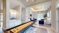 Located at 1500 German School Rd in Richmond, VA, this thoughtfully designed single-floor amenity space offers a well-rounded collection of rooms that balance comfort, productivity, and fitness. The living room serves as the centerpiece of the space, boasting soaring ceilings, large windows dressed with sheer curtains that flood the room with natural light, and rich wood-look flooring. A sleek white fireplace framed by built-in shelving anchors one wall, with a wall-mounted television above. Bold blue velvet lounge chairs and a shuffleboard table add a social, recreational atmosphere, while a wet bar with a beverage cooler and gold-tiled backsplash provides a stylish entertaining touch. A striking multi-arm chandelier illuminates the main gathering area. The exercise room is comprehensively outfitted with cardio machines, a multi-functional cable system, free weights, resistance equipment, and a large wall mirror, all set beneath an elegant coffered ceiling with ample natural light from surrounding windows. The dedicated office features a large conference table, ergonomic seating, comfortable lounge chairs, tall windows with dramatic navy drapes, and contemporary abstract artwork. Throughout, the space maintains a cohesive, upscale aesthetic with neutral tones, clean lines, and premium finishes.