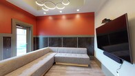 Located at 15 E Lane Ave in Columbus, Ohio, this single-floor amenity space combines modern design with functional versatility. The open-plan lounge area serves as the centerpiece, featuring light wood-look flooring, recessed lighting, and a distinctive coffered ceiling accent with a soft blue finish. A large wall-mounted flat-screen TV anchors the entertainment zone, complemented by a generous sectional sofa in neutral gray upholstery. A bold terracotta-orange accent wall with a dark wood panel wainscoting adds striking visual character, while an eye-catching circular ring chandelier overhead provides stylish ambient illumination. Adjacent to the lounge, a high-top bar counter with wood stools offers a casual seating alternative, ideal for informal gatherings or casual work sessions. The kitchenette is well-appointed with wood-grain cabinetry extending floor to ceiling, quartz countertops, a stainless steel sink, microwave, and dishwasher, all set against a clean white subway tile backsplash. A private meeting room provides a focused, quiet environment, furnished with a white rectangular table and a mix of pink and light blue ergonomic chairs beneath a drop ceiling with flush fluorescent lighting. The overall space presents a clean, contemporary aesthetic well-suited for community use or professional gatherings.