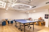 Located at 877 College St in Toronto's vibrant Bloordale neighbourhood, this single-floor recreation space is thoughtfully designed to encourage social interaction and active play. The room is finished with light hardwood flooring that brightens the interior and complements the clean white walls throughout.

At the heart of the space sits a full-size JOOLA ping pong table, offering ample room for competitive play. A foosball table anchors another zone of the room, while a pair of classic arcade machines add a retro gaming element to one corner. A dartboard mounted on the wall provides yet another activity option for visitors.

Casual seating, including yellow lounge chairs and low stools arranged around a round wooden table, creates a relaxed area for spectators or those taking a break between games. Industrial-style work and storage tables are positioned along the walls, adding functional surface space. A large built-in whiteboard with a warm wood trim spans one wall, offering a collaborative or instructional surface.

The ceiling features exposed white-painted pipes and ductwork alongside a circular pendant light fixture and recessed and track lighting, creating a modern industrial aesthetic while ensuring the space is well-lit. The overall ambiance is energetic yet welcoming, making this room well-suited for team-building activities, casual socializing, or recreational breaks.