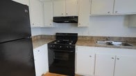 This single-story space includes a functional kitchen designed with efficiency in mind. The kitchen features white upper and lower cabinetry providing ample storage, complemented by granite-style laminate countertops that add a classic aesthetic. A black gas range with four burners serves as the focal point of the cooking area, paired with a matching black overhead vent hood for ventilation. A stainless steel sink is positioned nearby, offering a practical layout for food preparation and cleanup. The cabinetry is fitted with simple round knobs, maintaining a clean and understated look. Light hardwood flooring runs throughout, adding warmth to the space. The white walls and cabinetry create a bright, open feel, while the contrasting black appliances provide a bold visual element. The L-shaped counter configuration maximizes workspace and allows for smooth movement within the kitchen. Overall, the space presents a straightforward, no-frills kitchen layout well-suited for everyday cooking needs.