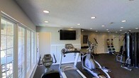 This fitness center is designed to accommodate a variety of exercise needs within a single, open-plan space. The room features a textured ceiling with recessed lighting, providing bright, even illumination throughout. Large windows along one wall allow natural light to filter in, creating an inviting atmosphere conducive to working out.

The cardio section is well-stocked, including treadmills, an elliptical trainer, and a stationary bike, all positioned to face a wall-mounted flat-screen television—ideal for entertainment during longer sessions. A wall clock is also mounted nearby for convenient time tracking.

Along the back wall, a dedicated free weights area offers a rack of dumbbells in varying sizes, catering to both beginner and advanced users. Additional strength and cardio equipment, including a stair climber, occupies the far end of the room, making effective use of the available floor space.

The flooring consists of dark, rubberized material suitable for high-impact activity, while the neutral-toned walls and white wainscoting contribute to a clean, organized aesthetic. A secondary door provides convenient access, and a bulletin board on the wall offers space for fitness information or community postings.