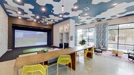 This single-floor amenity space combines bold design with versatile functionality. The most striking architectural feature is the ceiling, adorned with a large-scale navy and white hexagonal pattern, complemented by industrial-style pendant lighting and recessed fixtures that create a dynamic visual focal point throughout the space.

The main lounge area features polished concrete floors and exposed light brick walls that add texture and an urban, industrial character. Large black-framed windows flood the interior with natural light and offer views of the surrounding streetscape, while botanical-print curtains add a lively decorative contrast.

A built-in golf simulator anchors one end of the room, complete with a projected screen, turf mat, and dedicated display shelving showcasing trophies and golf equipment above navy cabinetry. A long communal wooden bar-height table with vibrant yellow lattice chairs serves as a central gathering and co-working surface, with an additional wall-mounted TV nearby.

A quieter lounge nook features a plush blue velvet sofa, mustard accent chair, wall-mounted television, and a geometric area rug, providing a more intimate retreat. A navy credenza with a beverage cooler adds convenience. Together, these elements create a lively, well-appointed social environment designed for both active recreation and casual gathering.