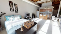 This open-concept studio showcases a compelling blend of industrial and contemporary design elements. The most striking architectural feature is the exposed wood beam ceiling, which adds warmth and character to the space while contributing to a sense of generous vertical volume. Sunlight streams in through the windows, casting bright natural light across the carpeted living and sleeping area, enhancing the airy, open ambiance.

The living space is thoughtfully arranged, with a comfortable bed, media console, and decorative accents — including a vintage trunk and wooden candleholders — that lend a curated, eclectic feel. A decorative wrought-iron wall piece adds an artistic touch to the neutral gray walls.

The kitchen is seamlessly integrated into the open floor plan, outfitted with warm wood-toned cabinetry, a tile backsplash, full-sized appliances including a refrigerator and microwave, and track lighting overhead. A peninsula with a butcher-block-style countertop and modern bar stools serves as both a functional prep area and a casual dining space, creating a natural transition between the kitchen and the living area. The overall design balances rustic and modern influences, resulting in a cohesive, inviting single-floor living environment.