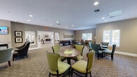 Located at 5303 Rue De Ville in Indianapolis, Indiana, this single-floor community living space offers a thoughtfully designed and multifunctional layout ideal for socializing and recreation. The interior is finished with warm taupe walls, white trim, and a patterned carpet that lends a cohesive, welcoming character throughout.

The lounge area is furnished with upholstered accent chairs, a settee, and a round coffee table, creating an intimate seating arrangement well-suited for conversation or relaxation. Wall-mounted televisions and framed artwork add visual interest, while recessed lighting provides a clean, well-lit ambiance. Large windows allow natural light to filter in and offer views of the outdoor pool area.

A game zone features a full-length shuffleboard table alongside a round dining table with cushioned chairs, providing a dedicated space for recreational activities and casual gatherings. This area connects fluidly with the rest of the space, encouraging easy movement and interaction.

The kitchenette area is equipped with rich dark wood cabinetry, a microwave, a small refrigerator, and counter space, making it practical for hosting events. Adjacent computer workstations and additional dining tables with colorful seating complete the space, supporting both productivity and community engagement. Together, these zones form a versatile and inviting communal environment.