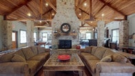 This single-story clubhouse delivers an impressive rustic aesthetic anchored by a dramatic floor-to-ceiling stone fireplace that serves as the focal point of the main gathering area. The soaring vaulted ceiling is framed by an intricate network of exposed timber beams and trusses, crafted from richly toned wood that adds warmth and architectural character to the entire space. Recessed lighting and pendant fixtures illuminate the room, complementing the natural light that filters in through multiple windows. The main lounge area is furnished with generous upholstered sofas arranged around a substantial rustic wooden coffee table, creating a welcoming and relaxed atmosphere ideal for socializing. A decorative millstone-style mirror adorns the stone fireplace mantel, reinforcing the space's refined yet rugged character. Beyond the main lounge, a billiard area is visible to one side, while a bar or kitchen area with dark cabinetry and bar seating is accessible on the other, making this clubhouse well-equipped for entertaining. Rich hardwood flooring ties all the areas together seamlessly. The overall ambiance is one of comfortable, lodge-inspired sophistication — a space designed for gathering, recreation, and relaxation.