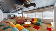 Located at 200 N Harvey Ave in downtown Oklahoma City, this single-floor communal space offers a lively and welcoming environment designed for collaboration and relaxation. The open layout is anchored by a bold, eclectic design aesthetic, highlighted by a multicolored carpet tile floor featuring vivid patches of red, orange, teal, and yellow that energize the entire space.

The main lounge area is furnished with an assortment of colorful seating — including patterned sofas, modular chairs, and bar-height tables with bright orange chairs — creating distinct zones for lounging, socializing, and casual dining. A wall-mounted flat-screen television adds an entertainment focal point, while framed artwork contributes to the creative atmosphere.

The exposed industrial ceiling, painted dark and fitted with track lighting and large HVAC ductwork, lends the space an urban, loft-like character. A row of large windows along one wall floods the interior with natural light and offers views of the surrounding cityscape.

A dedicated kitchen area supports the communal functions of the space, equipped with granite-style countertops, white cabinetry, a full refrigerator, microwave, sink, and a beverage station. The kitchen connects seamlessly to the lounge, making the overall space well-suited for gatherings, breaks, or community use.