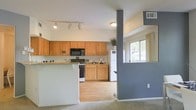 This single-floor residence offers a thoughtfully designed open-concept layout that seamlessly connects the kitchen and living areas. The kitchen is a functional and inviting space, featuring warm honey-toned wood cabinetry that extends to the upper walls, providing generous storage. Granite-style countertops add a touch of elegance, while the kitchen is equipped with black appliances including a microwave, range, and a full-size white refrigerator. A breakfast bar with countertop seating offers a casual dining option and serves as a natural divider between the kitchen and the adjacent living space. Track lighting overhead ensures the kitchen is well-lit, complementing the natural light that filters in through a window nearby. A distinctive gray accent column adds an architectural element that subtly separates the spaces while maintaining the open feel. The living area transitions smoothly from hardwood-style flooring in the kitchen to plush carpeting, creating distinct yet connected zones. The overall ambiance is bright and welcoming, with neutral tones and practical finishes that offer a comfortable, move-in-ready environment.
