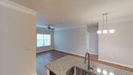 This single-story residence showcases an open-concept design that visually connects the kitchen and living areas, creating a cohesive and functional layout. The kitchen is appointed with granite countertops featuring a speckled finish, a stainless steel undermount sink with a modern pull-down faucet, and pendant lighting that adds a refined touch overhead. Crown molding trims the ceilings throughout, lending an elevated architectural detail to the space.

The living area extends naturally from the kitchen, finished with rich hardwood-style flooring that runs consistently across the main living spaces. A ceiling fan is centrally positioned, contributing both to the room's functionality and its overall aesthetic. Natural light enters through a window fitted with horizontal blinds, brightening the space during the day. Neutral-toned walls provide a clean, versatile backdrop throughout. A doorway visible in the background suggests additional rooms beyond the main living area, hinting at a well-organized floor plan. The overall ambiance is clean, contemporary, and practical.
