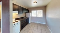 This single-floor unit offers a practical and straightforward layout with consistent light-toned ceramic tile flooring throughout. The kitchen is equipped with dark espresso-stained cabinetry paired with dark granite-style countertops, providing a notable contrast against the neutral beige walls. A white electric range, white refrigerator, and an under-cabinet range hood complete the kitchen's functional setup. A sliding window above the sink area allows natural light to filter in, offering views of surrounding greenery.

The living area is generously proportioned, featuring a textured popcorn ceiling, baseboard heating units along the walls, and a distinctive burgundy accent beam that adds a subtle architectural detail. The space is bright and open, well-suited for various furniture arrangements.

Adjacent to the main living area, a room features mirrored sliding closet doors that reflect light and add a sense of depth to the space. A wall-mounted air conditioning unit and a secondary window provide both ventilation and natural light. The hallway connects to additional rooms, each with tile flooring, baseboard heating, and windows allowing natural light throughout. The bathroom corridor reveals dark cabinetry consistent with the kitchen's design aesthetic.