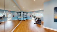 Located at 6158 Beacon Isles Dr in Town N' Country, FL, this dedicated fitness studio offers a thoughtfully designed space for a wide range of workout activities. The single-floor layout is finished with warm hardwood-style flooring throughout, providing a clean and durable surface ideal for exercise.

The main workout area is lined with full floor-to-ceiling mirrored walls, creating a sense of expansive space while allowing users to monitor their form during training. A ballet barre runs the length of the mirrored wall, making the studio well-suited for barre, dance, and stretching routines. Recessed lighting illuminates the space evenly from above, complemented by natural light streaming in through multiple windows that offer views of the surrounding outdoor greenery.

The studio is equipped with Echelon smart cycling bikes featuring integrated display screens, perfect for guided cardio sessions. Freestanding weight racks stocked with medicine balls and dumbbells provide strength training options, while wall-mounted smart fitness mirrors offer interactive workout programming. Motivational tropical-themed artwork adorns the light blue walls, reinforcing an energizing yet calming atmosphere. A wall-mounted HVAC unit ensures comfortable climate control year-round. With its versatile layout and comprehensive equipment, this fitness studio is designed to support a full spectrum of wellness activities.