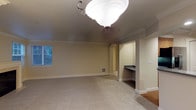 This single-floor space offers a clean, well-appointed layout with thoughtful architectural details throughout. The main room is generously sized, featuring plush wall-to-wall carpeting that adds warmth underfoot and neutral-toned walls that create a calm, inviting atmosphere. Crown molding runs along the ceiling perimeter, lending a refined, classic touch to the space. A decorative flush-mount ceiling light fixture serves as a subtle focal point overhead, providing ambient illumination throughout the room. A window along one wall allows natural light to filter in, offering a glimpse of lush greenery outside. A graceful arched passageway leads to a built-in alcove area, complete with a dark countertop desk surface — an ideal nook for a workstation or study area. The transition from carpet to hardwood flooring near the entryway adds visual contrast and hints at varied functional zones within the space. Recessed vents and well-placed light switches reflect practical, thoughtful construction. The overall ambiance is neutral, move-in ready, and versatile, making this space well-suited for a variety of uses and interior design styles.