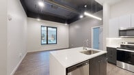 Located at 4201 S Navajo St in Englewood, Colorado, this contemporary single-floor residence offers a thoughtfully designed living experience with a distinct industrial-modern aesthetic. The open-concept living and kitchen area is anchored by a striking kitchen island with white quartz countertops and a stainless steel sink, complemented by a two-tone cabinetry scheme featuring dark wood lower cabinets and high-gloss white upper cabinets. Stainless steel appliances, including a refrigerator, range, microwave, and dishwasher, lend a polished, professional feel. An exposed duct ceiling finished in matte black adds an urban industrial character to the living area, which features wood-style flooring and windows overlooking the courtyard. A dedicated in-unit laundry area with a stacked washer and dryer adds everyday convenience. The bedrooms are carpeted and bright, with large windows framing views of the surrounding building courtyard. Two well-appointed bathrooms complete the unit — one featuring a sleek walk-in glass shower with subway tile surround and a dark wood vanity with a lit mirror, and the other offering a full soaking tub with a tiled surround. The overall design balances clean, contemporary lines with warm material choices throughout.