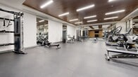 Located at 140 Ferguson Dr in Woodstock, Ontario, this fitness facility offers a thoughtfully designed exercise space spread across a single floor. The room is finished with a warm wood-paneled ceiling that contrasts elegantly with the crisp white textured walls, creating a polished and contemporary aesthetic throughout.

The cardio zone is equipped with treadmills, elliptical trainers, stationary bikes, and a rowing machine, all arranged to allow comfortable use and easy movement between equipment. Large mirrored panels line portions of the walls, visually expanding the space and allowing users to monitor their form during workouts. Integrated glass partitions further enhance the open, airy feel of the facility.

The strength training area is well-appointed with a multi-functional cable machine, a selection of dumbbells neatly racked along the wall, adjustable benches, and stability balls. A wall-mounted flat-screen television is positioned for visibility across the workout floor, adding to the overall user experience.

Bright recessed linear lighting runs across the ceiling, ensuring the entire space is evenly and thoroughly illuminated. The durable, smooth grey flooring is designed to withstand heavy equipment and high-traffic use. Overall, this fitness facility presents a clean, functional, and professionally outfitted environment suitable for a full range of workout routines.