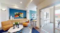 This thoughtfully designed single-floor communal space blends comfort and functionality through a series of distinct yet interconnected zones. The entrance opens into a welcoming lounge area anchored by a bold teal accent wall adorned with a large, colorful abstract canvas. Cognac leather lounge chairs surround a marble-topped coffee table set atop a geometric blue area rug, creating an inviting space for relaxation or casual conversation. Built-in cabinetry in warm wood tones lines one wall, adding both storage and warmth to the area. Framed photography, including a striking image of a red sculptural landmark, punctuates the white walls throughout.

Adjacent to the lounge, a dedicated co-working area features long, minimalist white desks running along both walls beneath bold teal paint, with pendant lighting hanging overhead and a floor-to-ceiling window at the far end that floods the space with natural light and outdoor views.

A separate conference room offers a more private setting, accommodating a full dining-style table and chairs beneath a sculptural gold chandelier. Floor-to-ceiling windows on two sides bathe the room in daylight, while a dramatic diagonal red accent wall adds a dynamic visual element. Patterned carpet with red accents ties the room's color palette together, making it equally suited for focused meetings or collaborative gatherings.