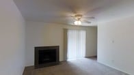 This single-floor residence offers a comfortable and functional layout designed for everyday living. The main living area serves as the focal point of the home, featuring a classic tile-surround fireplace that adds warmth and character to the room. Neutral carpet flooring runs throughout, complementing the light-toned walls and creating a clean, understated aesthetic. A ceiling fan with an integrated light fixture is centrally mounted, providing both air circulation and ambient lighting. A sliding glass door fitted with vertical blinds invites an abundance of natural light into the living space while offering access to the outdoors. The overall ambiance is calm and understated, with a simple, practical design that provides a solid foundation for personalization. The single-floor layout ensures easy accessibility and a seamless flow throughout the home, making it a straightforward and livable space suited to a range of lifestyles.