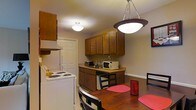 This single-floor space presents a practical and efficiently designed layout centered around a combined kitchen and dining area. The kitchen is outfitted with rich, dark wood upper and lower cabinetry that provides ample storage, complemented by a white electric range and a countertop featuring a microwave and toaster oven. The workspace is compact yet functional, making the most of the available footprint. Overhead, a flush-mount ceiling light provides steady ambient illumination throughout the kitchen area. The adjoining dining space is anchored by a warm-toned wooden dining table set with red placemats, lending a pop of color to the neutral interior. A distinctive pendant light with a dark-rimmed bowl shade hangs above the dining table, adding a focal point and directional warmth to the area. The flooring transitions subtly between the two zones, reinforcing the distinction between cooking and dining spaces. Neutral-toned walls and standard-height ceilings contribute to a grounded, understated ambiance throughout. A paneled interior door is visible leading to an adjacent area, suggesting additional rooms beyond the main living and kitchen zone. Overall, the space is straightforward and practical in its design.