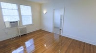 This single-floor residential space offers a thoughtfully laid-out interior defined by its warm, polished hardwood floors that reflect natural light throughout. The main living area is bright and open, featuring a large window fitted with blinds that allows ample daylight to fill the room. A classic cast-iron radiator along the wall speaks to the building's character and provides efficient radiant heating. A wall-mounted sconce adds a soft, ambient glow to the space. The crisp white walls and clean baseboards create a fresh, neutral backdrop that adapts well to a variety of furnishing styles. An open doorway connects the living area to an adjoining room, which appears to be a kitchen, where a white refrigerator is visible — suggesting a practical and accessible floor plan. The layout flows naturally between spaces, making the unit feel cohesive and functional. The well-maintained hardwood flooring runs consistently throughout, tying the rooms together and adding a timeless aesthetic quality. Overall, the space combines classic architectural details with a clean, move-in-ready condition, making it a practical and inviting single-floor residence.