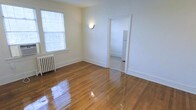 This single-floor residential space offers a thoughtfully laid-out interior defined by its warm, polished hardwood floors that reflect natural light throughout. The main living area is bright and open, featuring a large window fitted with blinds that allows ample daylight to fill the room. A classic cast-iron radiator along the wall speaks to the building's character and provides efficient radiant heating. A wall-mounted sconce adds a soft, ambient glow to the space. The crisp white walls and clean baseboards create a fresh, neutral backdrop that adapts well to a variety of furnishing styles. An open doorway connects the living area to an adjoining room, which appears to be a kitchen, where a white refrigerator is visible — suggesting a practical and accessible floor plan. The layout flows naturally between spaces, making the unit feel cohesive and functional. The well-maintained hardwood flooring runs consistently throughout, tying the rooms together and adding a timeless aesthetic quality. Overall, the space combines classic architectural details with a clean, move-in-ready condition, making it a practical and inviting single-floor residence.