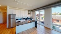 This single-floor space showcases a contemporary urban design characterized by clean lines, light hardwood flooring throughout, and a consistent neutral color palette. Exposed metallic ductwork runs along the ceilings, adding an industrial aesthetic that complements the modern finishes found throughout.

The kitchen is a standout feature, equipped with white cabinetry, dark granite countertops, stainless steel appliances including a range and refrigerator, a wall-mounted range hood, and a central island with matching dark countertop. Track lighting illuminates the space effectively, while a large floor-to-ceiling window floods the area with natural light and provides views of the surrounding neighborhood.

Additional rooms feature generous floor-to-ceiling windows and glass doors that bring in abundant natural daylight and offer outdoor views of the urban streetscape. The exposed ductwork is a recurring design element across these rooms, reinforcing the industrial-modern character of the unit.

The bathroom is well-appointed with a dark granite vanity top, white cabinetry with contemporary hardware, a large mirror, built-in shelving, tile flooring, and a combined tub and shower. The overall ambiance of the space is bright, open, and modern, with a cohesive design language maintained throughout.