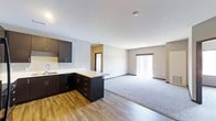 This single-floor space is designed with an open-concept layout that integrates the kitchen and living area into one cohesive, functional environment. The kitchen features dark espresso-toned cabinetry that extends along the wall and wraps into a peninsula, providing additional counter space and storage. Light-colored countertops offer a clean contrast against the dark cabinetry, and a sink is positioned along the perimeter wall for practical use. The kitchen flooring consists of wood-look planks in a warm tone, adding texture and visual warmth to the space.

The living area transitions seamlessly from the kitchen, distinguished by plush carpeting in a neutral gray tone. A sliding glass door at the far end of the living space allows natural light to fill the room and provides access to an outdoor area. The walls throughout are painted in a soft, neutral gray, complementing the overall modern and understated aesthetic. Recessed lighting in the ceiling provides even illumination across both the kitchen and living zones. A doorway visible in the background suggests additional rooms beyond the main open area, contributing to the overall functionality of the layout.