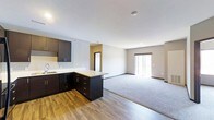 This single-floor space is designed with an open-concept layout that integrates the kitchen and living area into one cohesive, functional environment. The kitchen features dark espresso-toned cabinetry that extends along the wall and wraps into a peninsula, providing additional counter space and storage. Light-colored countertops offer a clean contrast against the dark cabinetry, and a sink is positioned along the perimeter wall for practical use. The kitchen flooring consists of wood-look planks in a warm tone, adding texture and visual warmth to the space.

The living area transitions seamlessly from the kitchen, distinguished by plush carpeting in a neutral gray tone. A sliding glass door at the far end of the living space allows natural light to fill the room and provides access to an outdoor area. The walls throughout are painted in a soft, neutral gray, complementing the overall modern and understated aesthetic. Recessed lighting in the ceiling provides even illumination across both the kitchen and living zones. A doorway visible in the background suggests additional rooms beyond the main open area, contributing to the overall functionality of the layout.