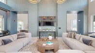 This thoughtfully designed single-floor amenity space offers an impressive blend of comfort, functionality, and contemporary coastal style. The living room is an immediate standout, anchored by soaring ceilings, soft sage-blue textured walls, and a centrally mounted flat-screen TV above an elegant wrought-iron console table. Paired globe pendant lights illuminate two plush sofas arranged around a substantial round wood coffee table, creating an inviting and balanced gathering space. Large windows throughout the space flood the interior with natural light and offer views of lush greenery and outdoor amenities. A built-in window bench seat, upholstered in rich blue fabric with fluted detailing and framed by floor-length curtains, provides a charming nook for relaxation. The kitchen and dining area features navy cabinetry with white quartz countertops, sage-green subway tile backsplash, a generous island with leather bar seating, and a built-in coffee station. A marble-topped dining table with a cushioned banquette completes the area. The game room includes a full-size pool table set against a fluted accent wall with a teal display shelf. Light wood-look tile flooring flows throughout, unifying each distinct zone into a cohesive, airy environment.