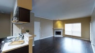 This single-floor space showcases an open-concept layout that seamlessly connects the kitchen and living area, creating a cohesive and functional floor plan. The living room is anchored by a classic corner fireplace with white tile surround, adding both a focal point and a cozy ambiance to the space. Dark hardwood flooring runs throughout, providing a rich contrast against the warm beige and tan walls accented with crisp white crown molding and baseboards.

Natural light filters generously through a large window fitted with horizontal blinds, brightening the living area and highlighting the clean lines of the interior. The kitchen area features a raised breakfast bar with light granite countertops, dark cabinetry, and a stainless steel appliance, partially visible from the main living space. The bar-style counter creates a natural division between the two zones while maintaining the open feel of the layout.

The neutral color palette, consistent flooring, and architectural details such as crown molding contribute to a polished, well-maintained interior. The overall ambiance is warm, inviting, and move-in ready, suited for comfortable everyday living.