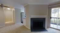 This single-floor residence offers a thoughtfully designed open-concept layout that seamlessly connects the living area to the kitchen. The living room serves as the heart of the home, anchored by a striking fireplace with a dark tile surround and a clean white mantel, providing a classic focal point against neutral-toned walls. Crown molding runs throughout, adding a refined architectural detail that elevates the overall aesthetic. The flooring transitions from plush, light-toned carpet in the living area to a smooth surface in the kitchen, clearly delineating the functional zones. The kitchen is equipped with stainless steel appliances, including a refrigerator and range, complemented by white upper cabinetry and a raised bar-height counter, making it both practical and inviting. A hallway visible from the living space leads to additional rooms, suggesting a well-organized floor plan. The neutral warm-beige color palette throughout creates a calm and welcoming ambiance, while the open sightlines between the living and kitchen areas make the space feel connected and functional. Crown molding, clean trim work, and consistent finishes contribute to a cohesive and well-maintained interior.