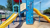 This outdoor playground is a lively, well-equipped recreational space designed to engage children of various ages and skill levels. The centerpiece of the space is a multi-element play structure constructed with bold blue steel framing, accented by red, yellow, and green components that create a visually stimulating environment. A prominent rock climbing wall with multicolored handholds provides a physical challenge for young climbers, while two bright yellow slides of varying configurations offer fast-paced fun. Decorative panel elements featuring cutout wave patterns and grid designs add visual interest to the structure's towers. The ground beneath the equipment is covered in wood chip surfacing, providing a cushioned, safety-conscious base. The playground is set within an open, sun-drenched area surrounded by mature trees that offer partial shade along the perimeter. Residential buildings are visible in the background, suggesting this space is situated within or adjacent to a neighborhood setting. The overall ambiance is cheerful and energetic, making it an inviting destination for active outdoor play in a community-oriented environment.
