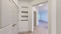 This single-floor residence is designed with a clean, contemporary aesthetic defined by crisp white walls, light wood-look flooring, and an open layout that promotes a seamless flow between living spaces. The entryway features built-in recessed wall shelving, offering convenient display or storage space, and opens into the main living area beyond an arched passageway.

The living room is spacious and airy, benefiting from a ceiling fan, direct outdoor access through a glass door, and large windows that frame views of mature trees, filling the space with natural light. Adjacent to the living area, the kitchen is well-appointed with light wood-toned cabinetry reaching toward the ceiling, granite countertops, a subway tile backsplash, a central island, and a full suite of stainless steel appliances including a refrigerator, range, and built-in microwave.

The bedroom features plush carpeting, dual windows overlooking greenery, and a ceiling fan, creating a comfortable and serene retreat. The bathroom is finished with granite countertops, wood-tone cabinetry, and a tiled tub-shower surround with a marble-like pattern. Light wood-look flooring runs consistently throughout the non-carpeted areas, unifying the overall design.
