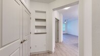 This single-floor residence is designed with a clean, contemporary aesthetic defined by crisp white walls, light wood-look flooring, and an open layout that promotes a seamless flow between living spaces. The entryway features built-in recessed wall shelving, offering convenient display or storage space, and opens into the main living area beyond an arched passageway.

The living room is spacious and airy, benefiting from a ceiling fan, direct outdoor access through a glass door, and large windows that frame views of mature trees, filling the space with natural light. Adjacent to the living area, the kitchen is well-appointed with light wood-toned cabinetry reaching toward the ceiling, granite countertops, a subway tile backsplash, a central island, and a full suite of stainless steel appliances including a refrigerator, range, and built-in microwave.

The bedroom features plush carpeting, dual windows overlooking greenery, and a ceiling fan, creating a comfortable and serene retreat. The bathroom is finished with granite countertops, wood-tone cabinetry, and a tiled tub-shower surround with a marble-like pattern. Light wood-look flooring runs consistently throughout the non-carpeted areas, unifying the overall design.