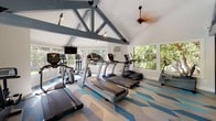 Located at 444-445 N Amelia Ave in San Dimas, California, this single-story fitness center is a well-appointed exercise facility designed to inspire an active lifestyle. The space is defined by its striking vaulted ceiling with bold gray exposed truss work, which lends an open, airy quality to the interior. A ceiling fan circulates air throughout, complementing the wall-mounted HVAC unit for year-round comfort.

The floor is covered in a vibrant geometric pattern in shades of blue, gray, and white, adding a dynamic and energetic visual element to the space. Large windows and glass French doors line the walls, flooding the interior with abundant natural light and offering pleasant views of the surrounding greenery.

The fitness area is equipped with multiple treadmills, elliptical machines, a stationary bike, and a stair climber, all from the True fitness brand. A wall-mounted flat-screen television provides entertainment during workouts. One wall features a bold, graphic fitness-themed mural with motivational typography, reinforcing the active atmosphere of the space.

A glass sliding door leads to an adjacent area with warm pendant lighting, visible from the main fitness floor. Built-in wooden cubby shelving provides convenient storage for personal belongings. The overall ambiance is bright, modern, and motivating, making this fitness center a functional and visually engaging community amenity.