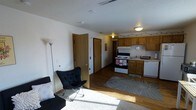 This single-floor space offers an efficient and functional open-concept layout that seamlessly integrates the living area and kitchen. The living space features plush carpeting near the seating area, which transitions into warm hardwood-style flooring that extends throughout the rest of the unit. A textured ceiling adds subtle visual interest overhead, while natural light filters in to brighten the interior.

The kitchen is well-appointed with medium-toned wood cabinetry that provides ample storage, a white electric range, a dishwasher, and a double-basin sink. The cabinetry layout allows for efficient meal preparation, and a decorative rug helps define the kitchen zone within the open floor plan.

A central hallway connects additional areas of the unit, with interior doors leading to private spaces. The layout flows naturally, making the space feel open and easy to navigate despite its compact footprint. Overhead lighting fixtures and a floor lamp in the living area ensure the space remains well-lit throughout. The overall ambiance is cozy and practical, with a clean, neutral palette that provides a versatile backdrop for a variety of furnishing styles. Ceiling vents indicate a central HVAC system for year-round comfort.