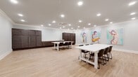 This single-floor space offers a well-designed, open-plan interior that balances functionality with a creative aesthetic. The main area is generously proportioned, accommodating multiple white farmhouse-style tables paired with dark chairs, making it well-suited for workshops, meetings, or collaborative events. Light wood-tone flooring runs throughout, lending warmth and continuity to the space.

Recessed lighting is installed across the ceiling in an abundant grid pattern, ensuring thorough, even illumination throughout. Crown molding trims the perimeter of the ceiling, adding a refined architectural detail. A ceiling-mounted HVAC unit provides climate control discreetly overhead.

Along one wall, a run of dark espresso-toned cabinetry provides substantial storage, complemented by a counter with a sink—ideal for catering or event preparation. A glass-paneled interior door connects to an adjacent room or corridor, adding visual depth and accessibility.

A striking gallery wall displays three large-scale figurative paintings in soft aqua tones with vivid pops of color, functioning as a bold focal point and giving the space a distinctly artistic character. The overall ambiance is clean, bright, and modern, with a creative energy suited to a wide range of professional or social gatherings.