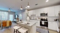 Located at 350 Bay Laurel Dr in Saint Augustine, FL, this one-bedroom, one-bathroom residence offers a thoughtfully designed single-floor layout with modern finishes throughout. The open-concept kitchen and living area serves as the heart of the home, featuring white shaker cabinetry, a white subway tile backsplash, granite countertops, and a full suite of stainless steel appliances. A central kitchen island with seating, illuminated by sleek gold pendant lights, provides an ideal space for casual dining and entertaining. Light wood-style flooring runs throughout the main living areas, complementing the neutral color palette. The living room is bright and inviting, benefiting from natural light through large windows and a glass door, creating an airy, open atmosphere. The bedroom is a comfortable retreat with plush carpet, a ceiling fan, and generous windows dressed with navy drapes that allow for natural light while maintaining privacy. A dedicated walk-in closet with wire shelving provides ample storage. The full bathroom is finished with large-format tile in the tub/shower surround, a granite-topped vanity with white cabinetry, and wood-style flooring, offering a clean and polished aesthetic throughout.
