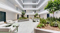 Located at 501 Broadway in Santa Monica, California, this interior courtyard serves as a central communal hub within a multi-story building. The space is defined by its open atrium architecture, with upper-level balconies and metal railings overlooking the ground floor, allowing natural light to filter down through a skylight above. The expansive floor is finished in large-format, light gray ceramic tiles that lend a clean, contemporary aesthetic throughout.

Generous raised concrete planters are strategically positioned across the courtyard, filled with a curated mix of lush greenery including snake plants, succulents, tropical shrubs, and mature trees, bringing a vibrant, biophilic quality to the interior environment. Wall-mounted sconce lighting accompanies each dark teal doorway along the perimeter, providing a warm, inviting glow.

Multiple distinct seating zones furnish the space for varied uses. A modular sectional sofa in neutral tones with a sage green frame anchors one area, complemented by a matching low coffee table. Nearby, a casual dining zone features round yellow tables with matching chairs, adding a pop of color and an informal gathering spot. A stainless steel elevator is accessible from the courtyard, ensuring convenient connectivity throughout the building. The overall ambiance is bright, airy, and meticulously designed to foster community and comfort.