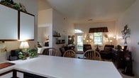 This single-floor home offers an open, interconnected layout that seamlessly blends the dining, living, and sleeping areas into a cohesive and comfortable living environment. The dining area features a clean, light-colored countertop or table in the foreground, surrounded by classic wooden chairs, creating a welcoming space for meals and gatherings. Beyond the dining area, the living room is furnished with plush, dark upholstered seating arranged around a flat-screen television, providing a relaxed setting for entertainment and lounging. The space is brightened by an arched front door flanked by windows and additional windows dressed with patterned valances, allowing generous natural light to fill the interior. Vaulted ceilings add a sense of openness and volume throughout. Decorative floating shelves display personal accents, contributing to the home's warm and lived-in character. A glimpse into the adjacent bedroom reveals a neatly made bed and soft lamp lighting, suggesting a calm and restful retreat. Overall, the home presents a cohesive, cozy aesthetic with a neutral color palette, warm wood tones, and thoughtful furnishings that make it feel both functional and inviting.