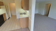 This single-floor residence offers a practical and comfortable layout designed for everyday living. The kitchen is equipped with warm wood-toned cabinetry, a double-basin stainless steel sink, and light-colored countertops that provide ample workspace. The kitchen flooring features classic square tile, adding durability to the space. A structural column subtly defines the boundary between the kitchen and the adjacent living area, creating a sense of separation while maintaining an open, connected feel. The living area is finished with neutral wall-to-wall carpeting, contributing to a warm and quiet atmosphere. Walls throughout are painted in soft, neutral tones, lending the space a clean and understated aesthetic that is easy to personalize. The layout is straightforward and efficient, with a smooth transition between the kitchen and the main living space. The overall design is practical and move-in ready, with consistent finishes that create a cohesive look throughout the home. The space is well-suited for those seeking a low-maintenance, functional residence with a simple, comfortable interior.