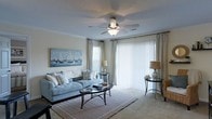 Located at 4912 Signature Dr in Myrtle Beach, SC, this single-story space embraces a coastal aesthetic throughout. The living area serves as the centerpiece, showcasing soft, neutral tones and plush carpeting underfoot. A ceiling fan with a light fixture provides both comfort and illumination, while crown molding along the ceiling adds a refined architectural touch.

Natural light flows generously into the room through a large sliding glass door dressed with vertical blinds, complemented by additional windows framed by elegant draping curtains. The space is furnished with a tufted, light blue sofa accented by decorative pillows, paired with a sleek oval glass coffee table and coordinating side tables. A nautical-themed wall canvas depicting sailboats reinforces the coastal character of the space.

The overall ambiance is calm and inviting, with a palette of soft blues, whites, and warm neutrals that evoke the relaxed atmosphere of beachside living. The thoughtful layout and design elements combine to create a comfortable, well-lit environment well-suited to the Myrtle Beach locale.