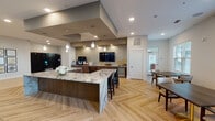 This thoughtfully designed single-floor communal space combines an open-plan kitchen, lounge, and dining areas into a cohesive, upscale environment. The kitchen area serves as a central hub, anchored by a generously sized island topped with dramatic veined granite. Dark walnut cabinetry, decorative pendant lighting, a mosaic tile backsplash, and built-in appliances — including a beverage refrigerator and coffee station — provide both function and visual interest. A wall-mounted television and a defined ceiling soffit add architectural structure overhead, while herringbone-patterned light wood flooring flows throughout, unifying the space.

The lounge area features soaring ceilings, creating an airy, open atmosphere. Curated seating arrangements — including plush swivel chairs, upholstered banquettes, and accent tables — are organized around wall-mounted televisions and complemented by abstract artwork. A textured stone accent wall adds warmth and depth to the built-in banquette nook.

A bright dining area positioned along windowed walls allows natural light to fill the space, with views toward an outdoor terrace accessible through floor-to-ceiling sliding glass doors framed by sheer drapery. Throughout, the design balances contemporary aesthetics with comfortable, functional furnishings suited for social gatherings.