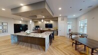 This thoughtfully designed single-floor communal space combines an open-plan kitchen, lounge, and dining areas into a cohesive, upscale environment. The kitchen area serves as a central hub, anchored by a generously sized island topped with dramatic veined granite. Dark walnut cabinetry, decorative pendant lighting, a mosaic tile backsplash, and built-in appliances — including a beverage refrigerator and coffee station — provide both function and visual interest. A wall-mounted television and a defined ceiling soffit add architectural structure overhead, while herringbone-patterned light wood flooring flows throughout, unifying the space.

The lounge area features soaring ceilings, creating an airy, open atmosphere. Curated seating arrangements — including plush swivel chairs, upholstered banquettes, and accent tables — are organized around wall-mounted televisions and complemented by abstract artwork. A textured stone accent wall adds warmth and depth to the built-in banquette nook.

A bright dining area positioned along windowed walls allows natural light to fill the space, with views toward an outdoor terrace accessible through floor-to-ceiling sliding glass doors framed by sheer drapery. Throughout, the design balances contemporary aesthetics with comfortable, functional furnishings suited for social gatherings.
